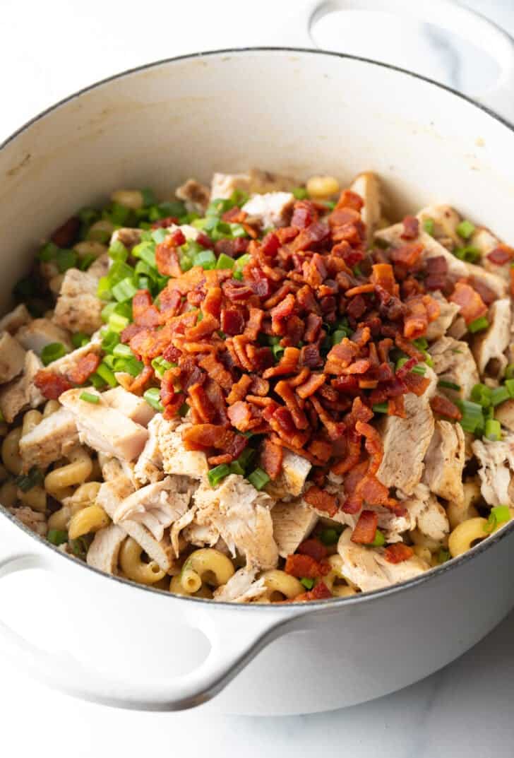 Adding chopped cooked bacon on top of the pasta and chicken in a cooking pot.