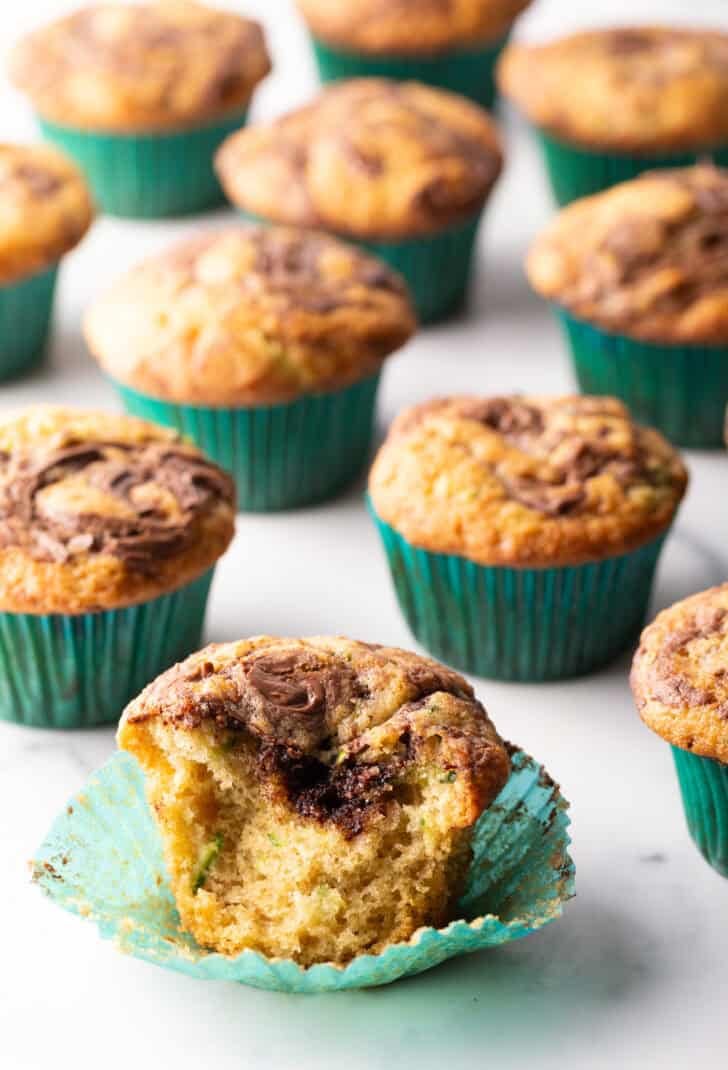 A dozen zucchini muffins with Nutella swirls, in green cupcake wrappers. One muffin has been split in half to show the inside.