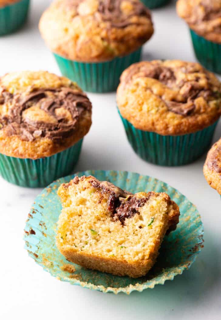A dozen zucchini muffins with Nutella swirls, in green cupcake wrappers. One muffin has been split in half to show the inside.