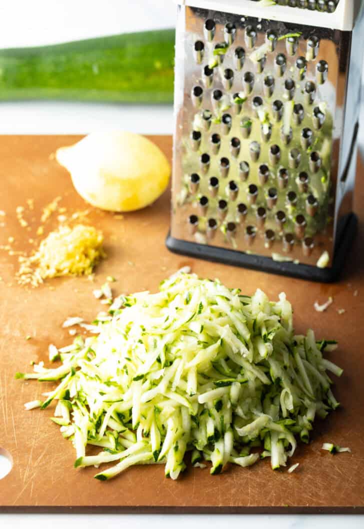 Box grater, whole lemon, and pile of shredded zucchini on a wood cutting board.