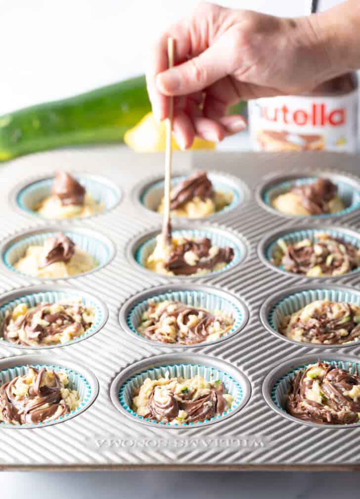 Hand using a wood skewer to swirl nutella into unbaked zucchini muffins in a baking tin.