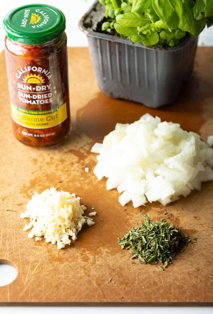 Recipe ingredients on a cutting board. Jar of sun-dried tomatoes, diced onion, minced garlic, fresh chopped herbs and a box of baby spinach leaves.