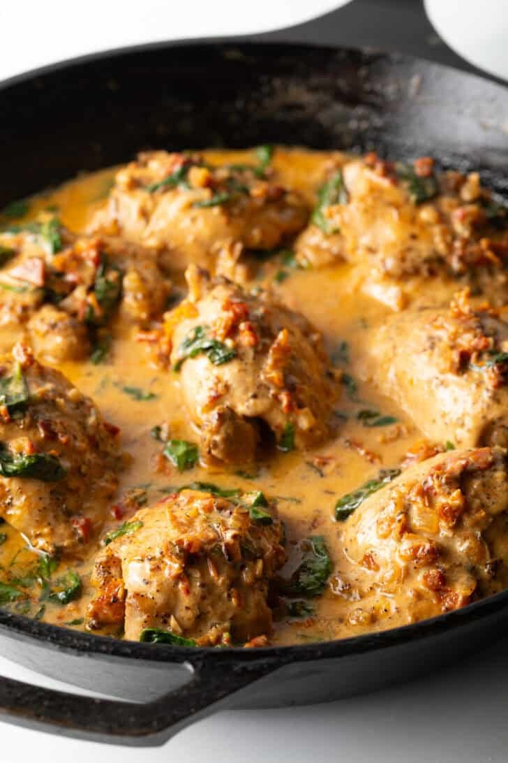 Chicken thighs simmering in a creamy Tuscan sauce in a cast iron skillet.