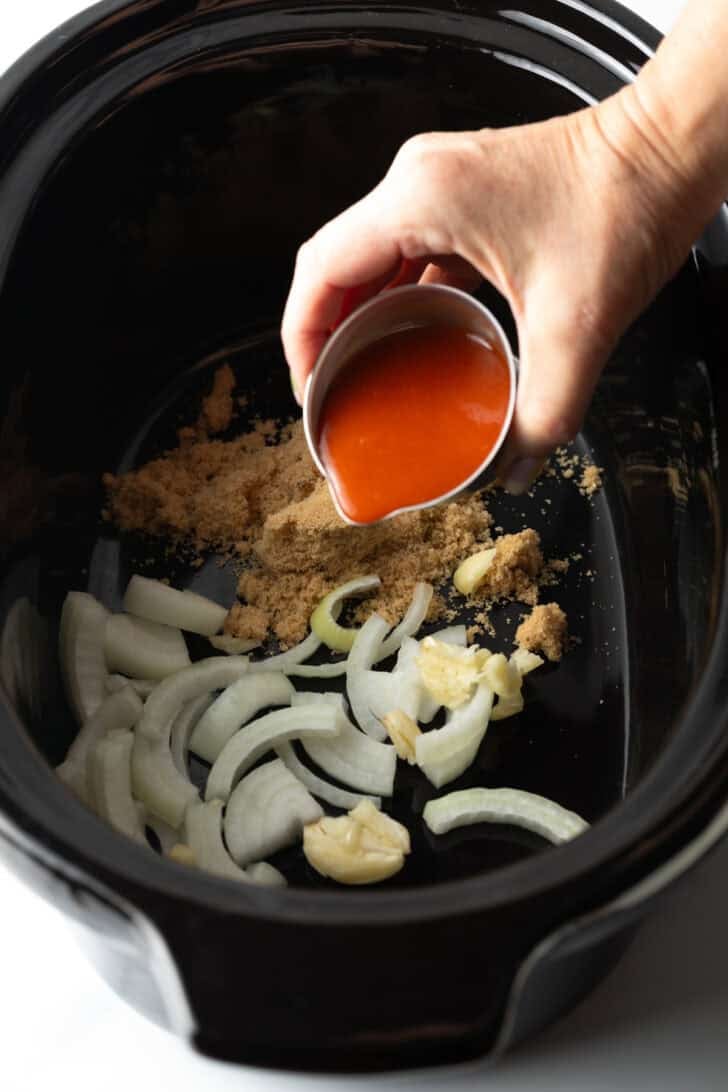 Pouring sauce from a small metal cup into the slow cooker with brown sugar and onions.