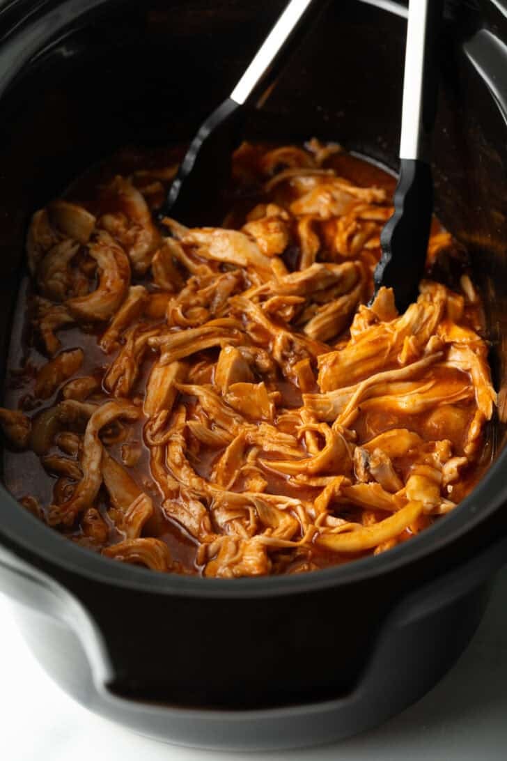 Tongs in a black crockpot with pulled bbq chicken.