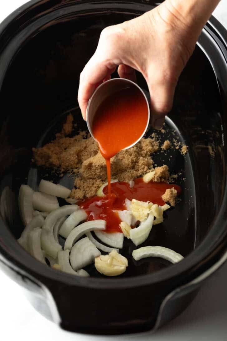Pouring sauce from a small metal cup into the slow cooker with brown sugar and onions.