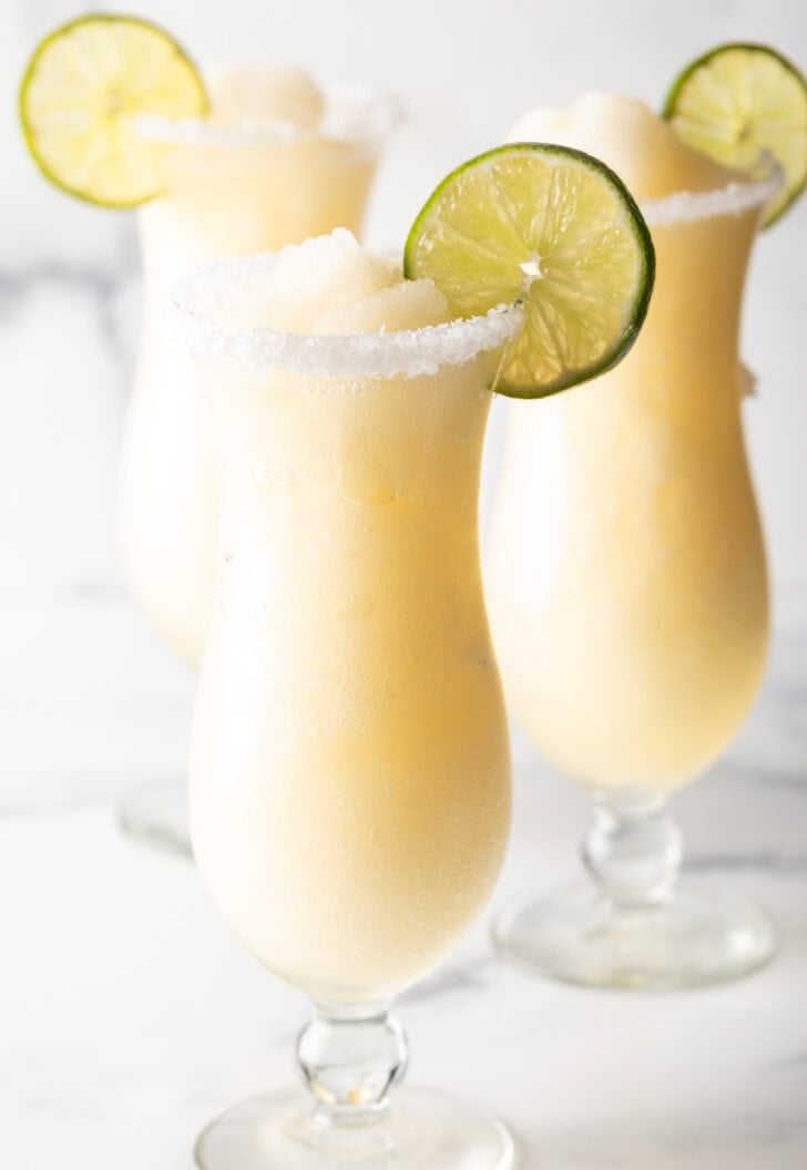 3 frozen margaritas in hurricane glasses, each garnished with a slice of lime and salt around the rim.