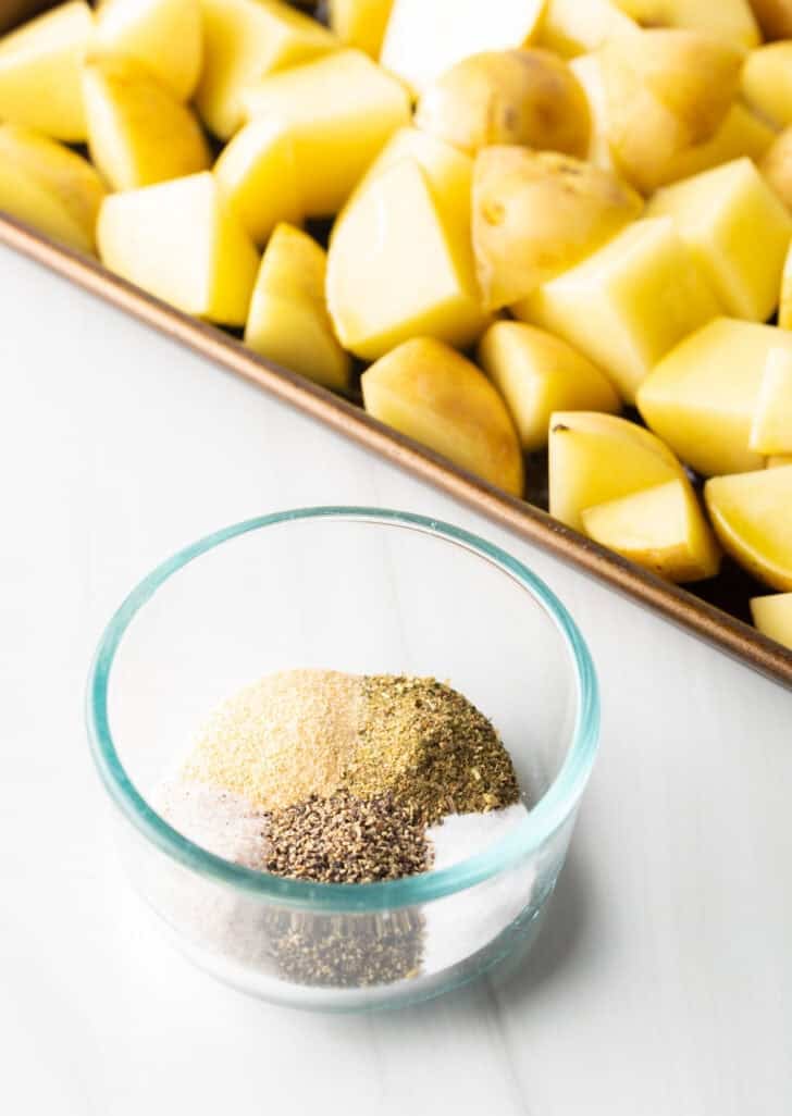 Raw pieces of yukon potatoes on a baking sheet, with a glass dish on the side filled with assorted spices.