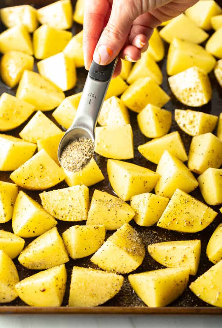 Hand sprinkling seasonings from a small teaspoon overtop of potatoes chunks on a baking sheet.