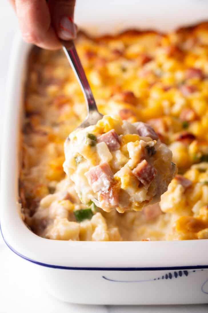 Hand with metal spoon full of ham and potato casserole, showing to camera.