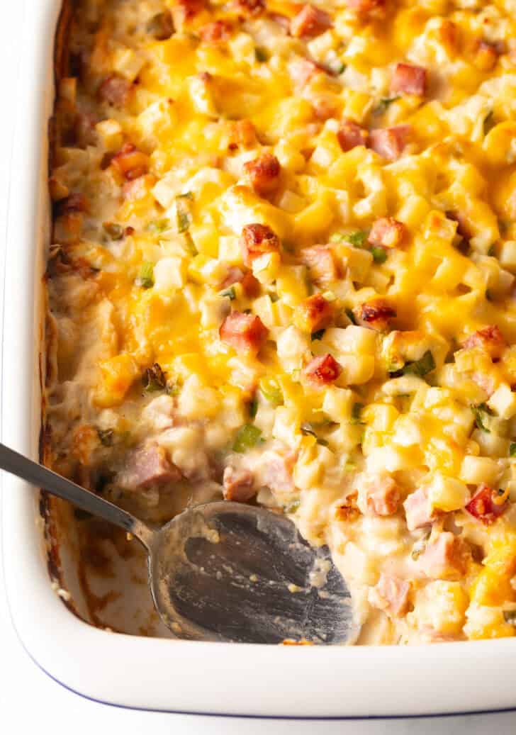 Top down view white casserole dish with ham and potatoes, and a spoon in the dish where a serving has been removed.