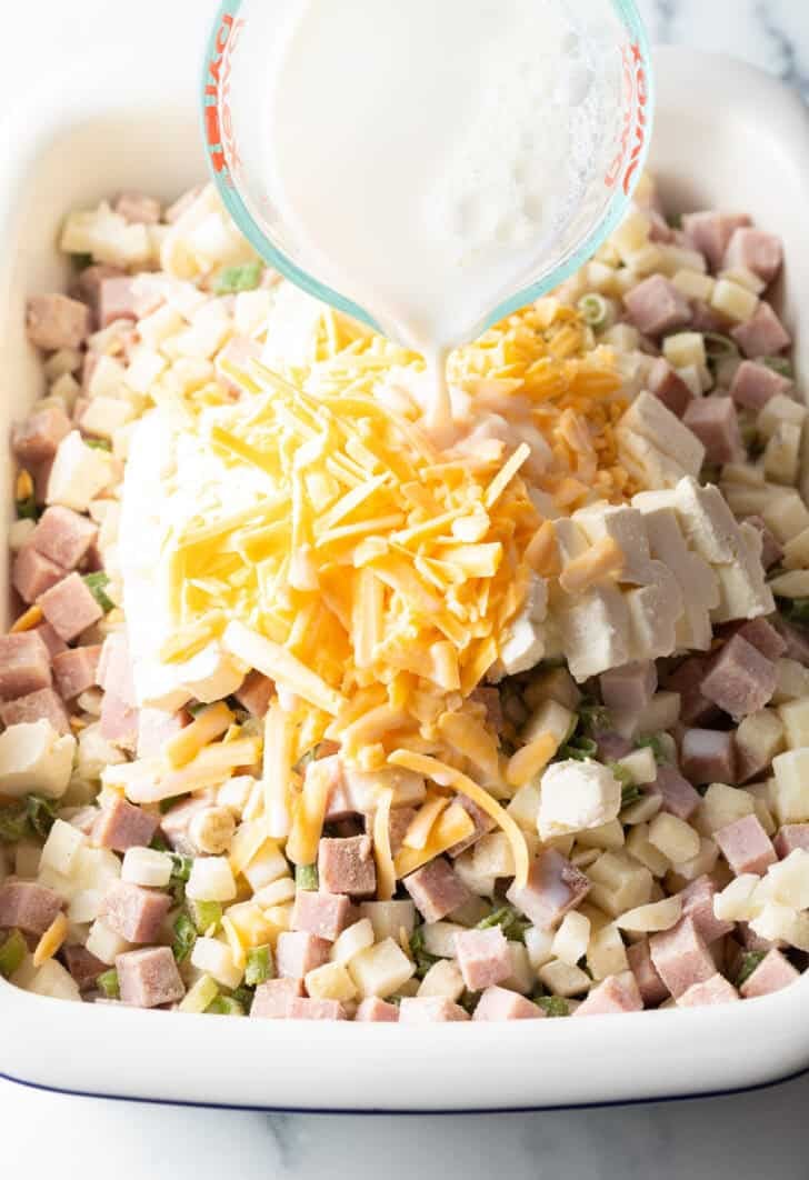 Adding milk to the chopped potatoes, ham, and shredded cheese in a casserole dish.