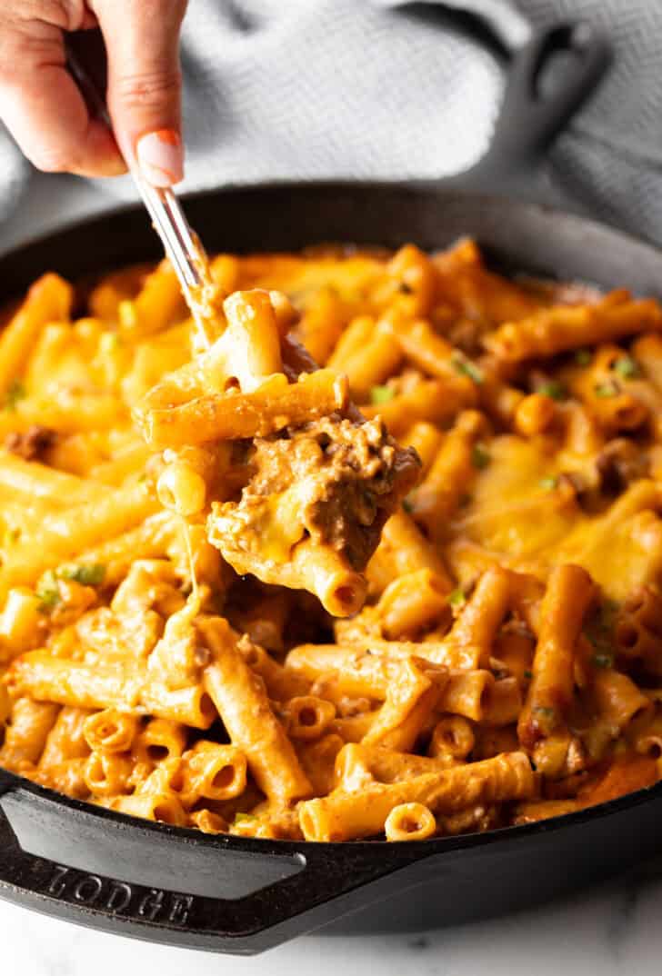 Hand using a large spoon to scoop noodle cheeseburger casserole from a large skillet towards camera.