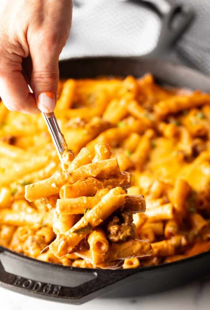 Hand using a large spoon to scoop noodle cheeseburger casserole from a large skillet towards camera.