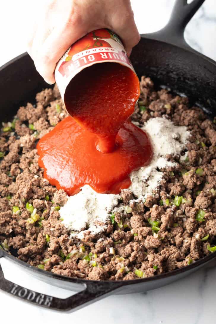 Hand adding canned tomato sauce to a skillet of cooked ground beef and ranch seasoning mix.