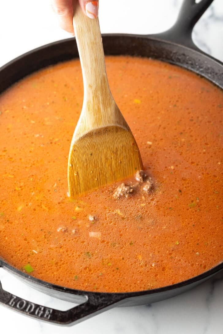 Using a wood spatula to stir tomato and beef mixture in a cast iron skillet.