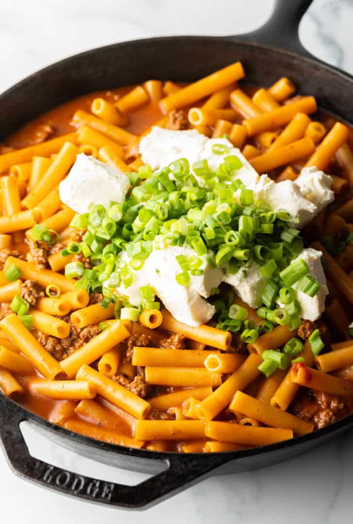 Adding cream cheese and chopped green onions to a cast iron skillet with ground beef and noodles.