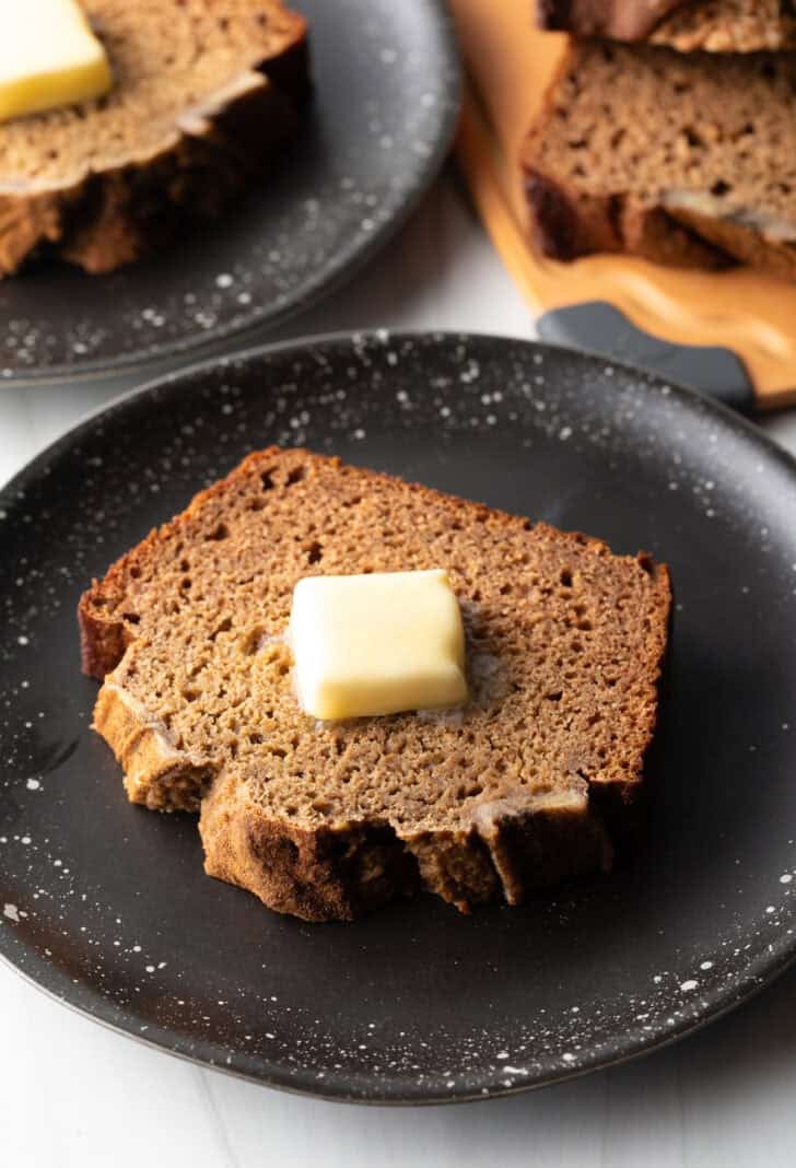 Thick slice of banana bread with a pat of butter on top, on a black plate.