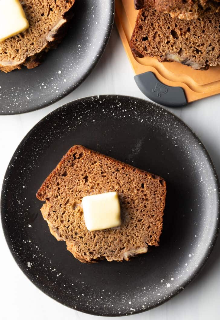 Thick slice of banana bread with a pat of butter on top, on a black plate.