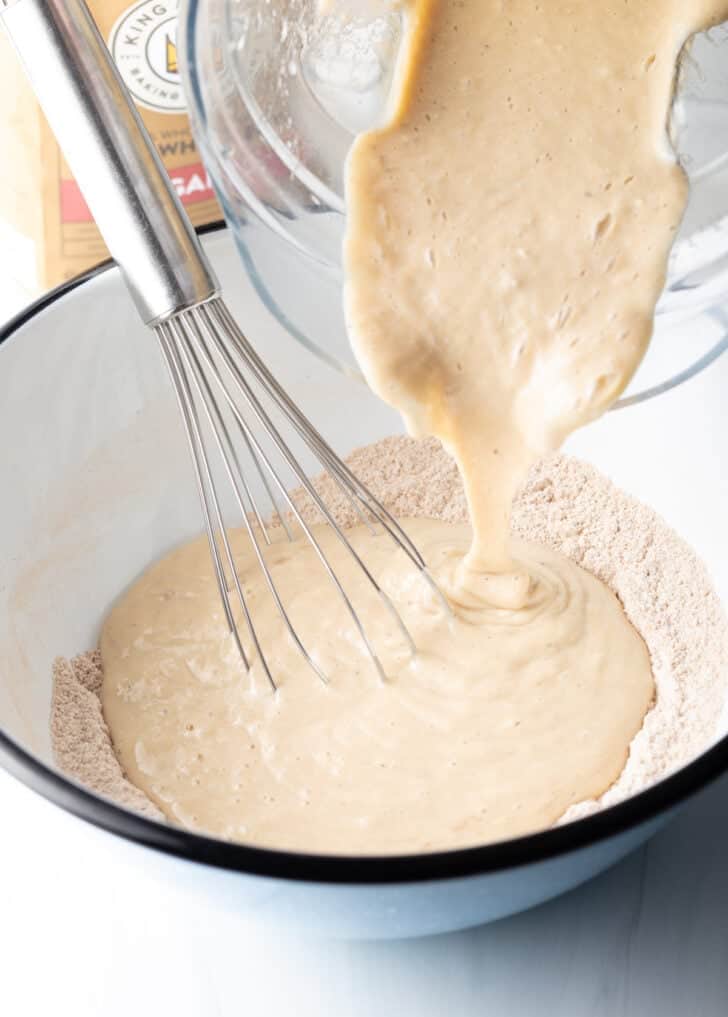 Adding wet ingredients from a glass bowl to dry ingredients in a large white mixing bowl.