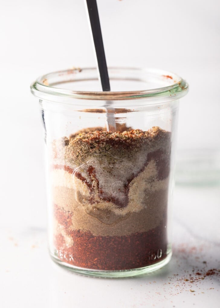 Spoon stirring layers of chicken taco spices in a tall glass jar.