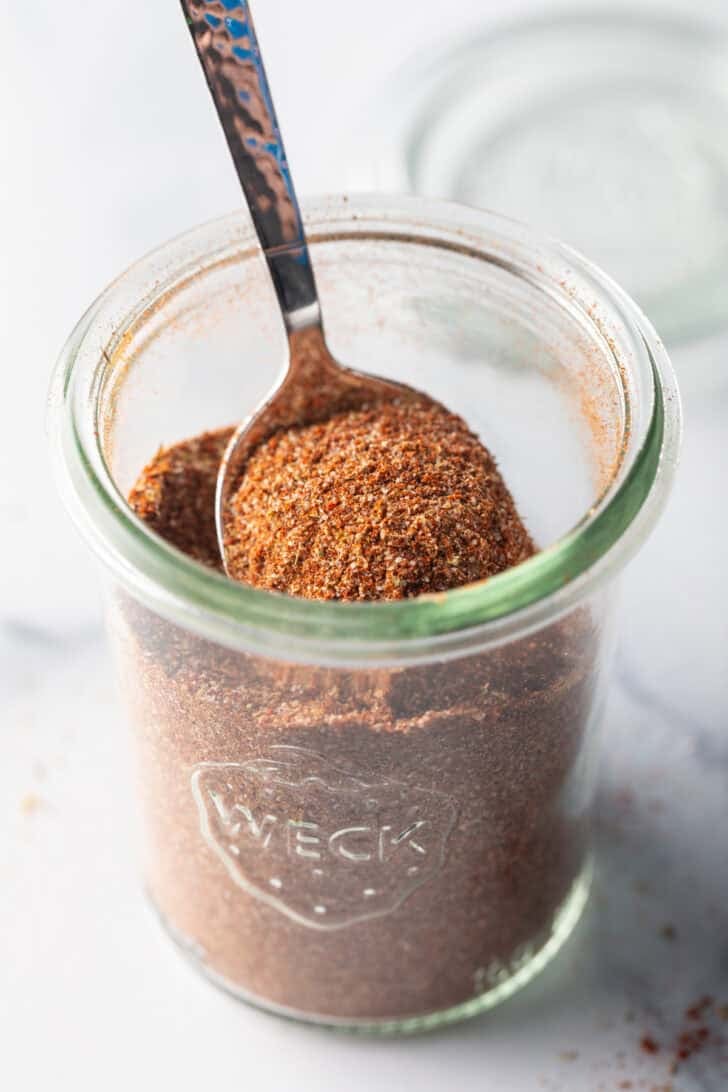 A spoon digging into a small glass jarful of chicken taco seasoning.