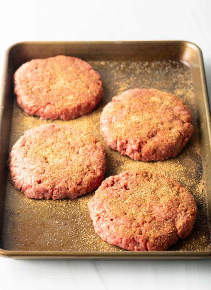 4 raw seasoned burger patties on a baking sheet.