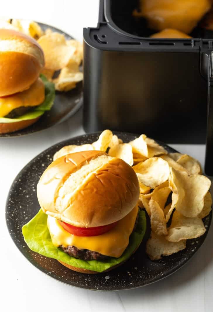 Aair fryer hamburger loaded with cheese, lettuce, tomatoes and pickle slices on a plate with potato chips.