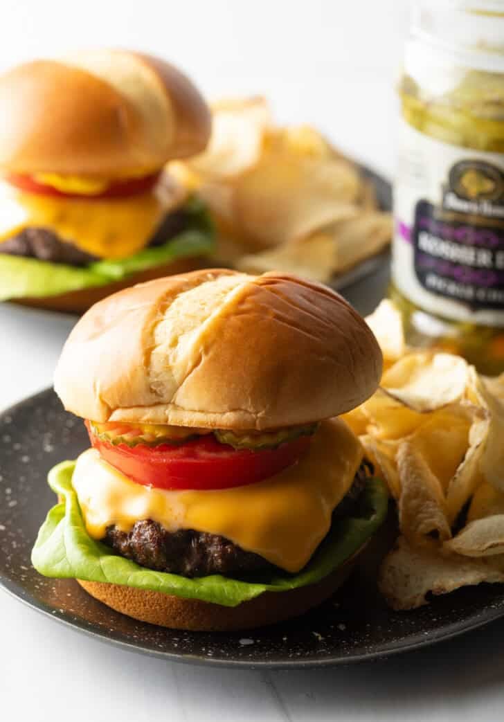 Aair fryer hamburger loaded with cheese, lettuce, tomatoes and pickle slices on a plate with potato chips.