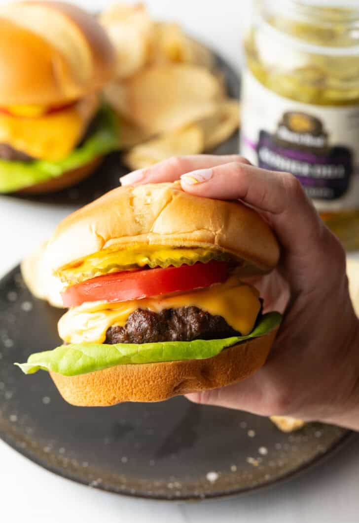 Hand holding a juicy air fryer hamburger loaded with cheese, lettuce, tomatoes and pickle slices.
