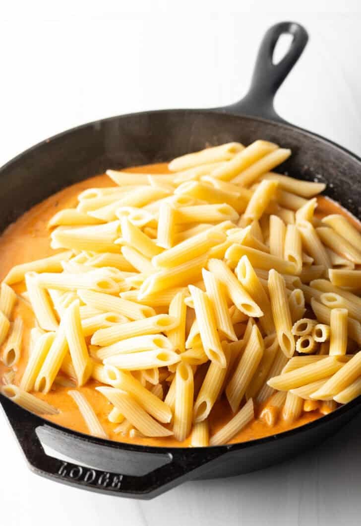 Adding penne noodles to the cream sauce in a cast iron skillet.