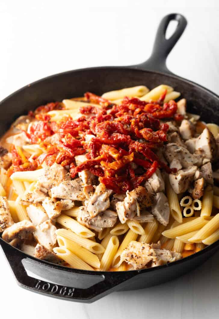 Adding sundried tomatoes to chopped chicken and penne pasta in a cast iron skillet.