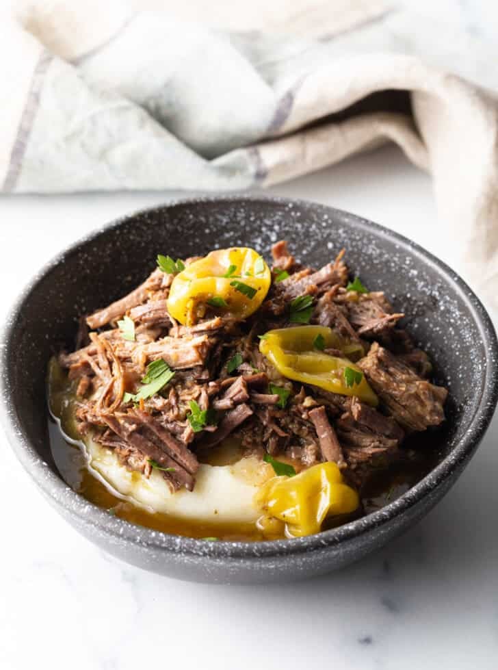 Black bowl with a layer of mashed potatoes topped with shredded pot roast and pepperoncinis.