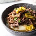 Black bowl with a layer of mashed potatoes topped with shredded pot roast and pepperoncinis.
