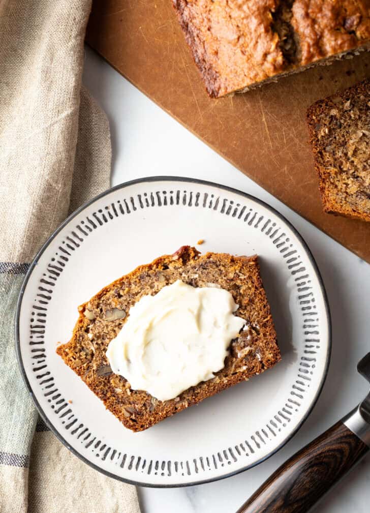 Top down view slice of banana bread on a white plate, butter is smeared across the slice.