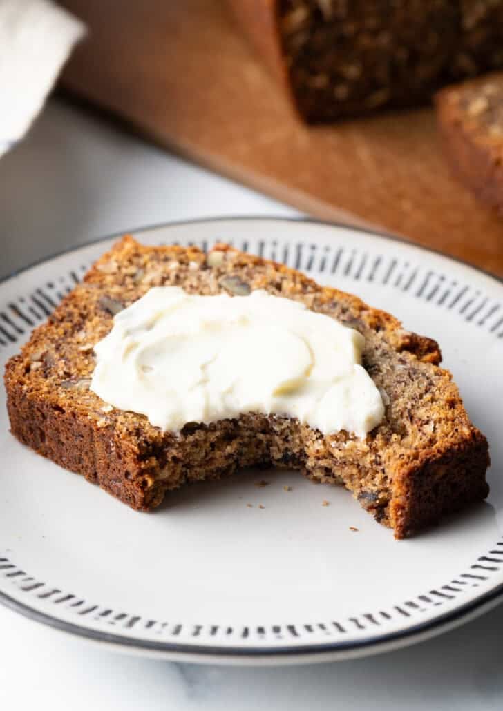 Slice of banana bread on a white plate, butter is smeared across the slice. A bite has been taken out of one corner.