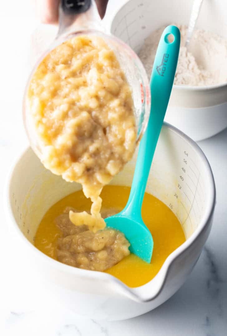 Adding mashed bananas from a measuring cup into a white mixing bowl with butter and a blue spatula.