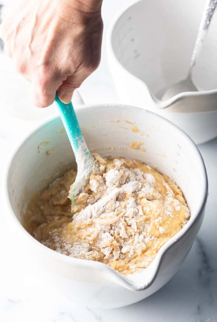 Hand using a blue spatula to stir banana bread batter in a large white measuring bowl.