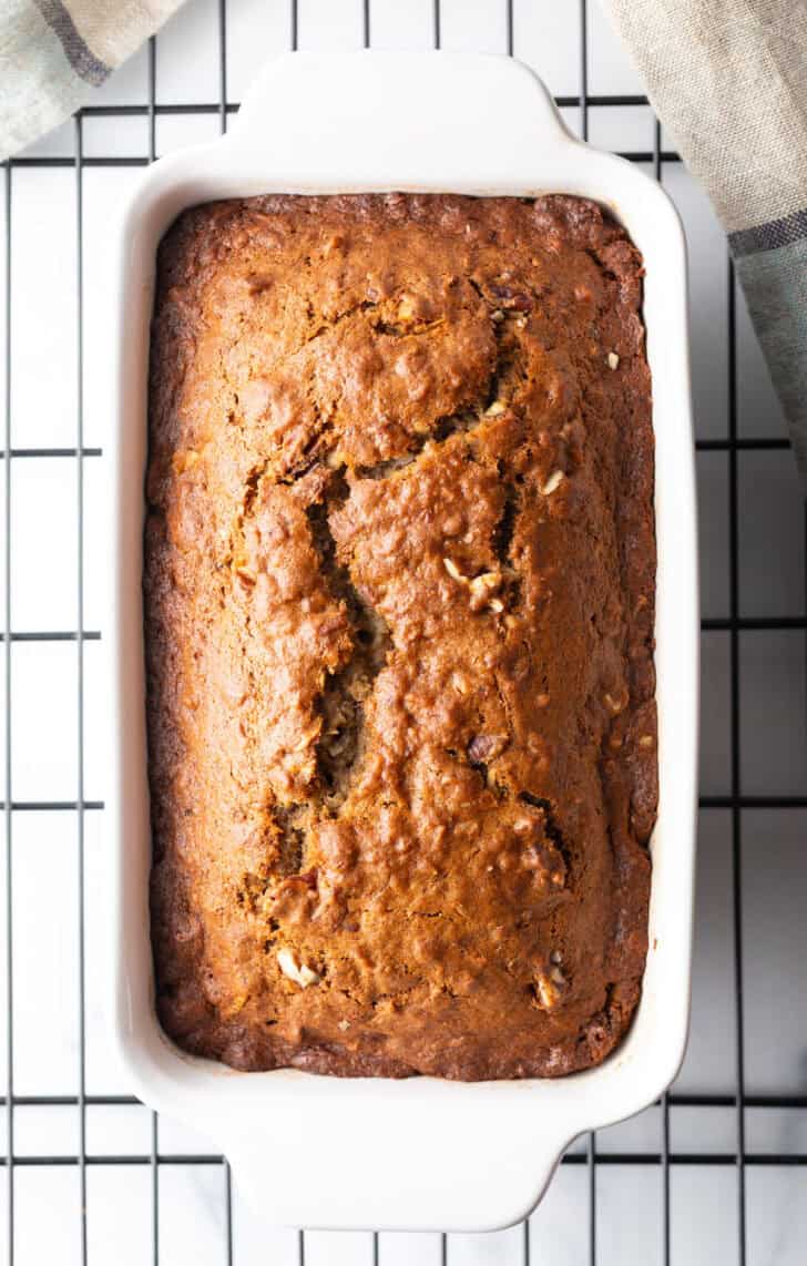 Top down view loaf of moist banana bread baked in a white loaf pan, cooling on a wire rack.