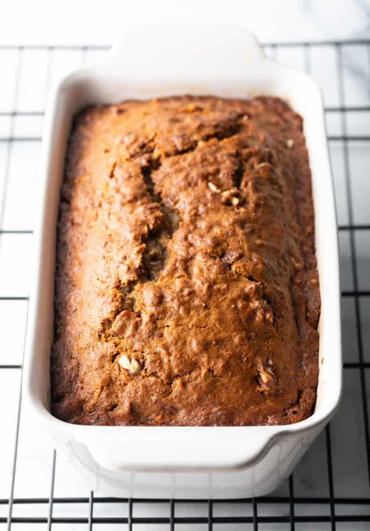 Loaf of moist banana bread baked in a white loaf pan, cooling on a wire rack.