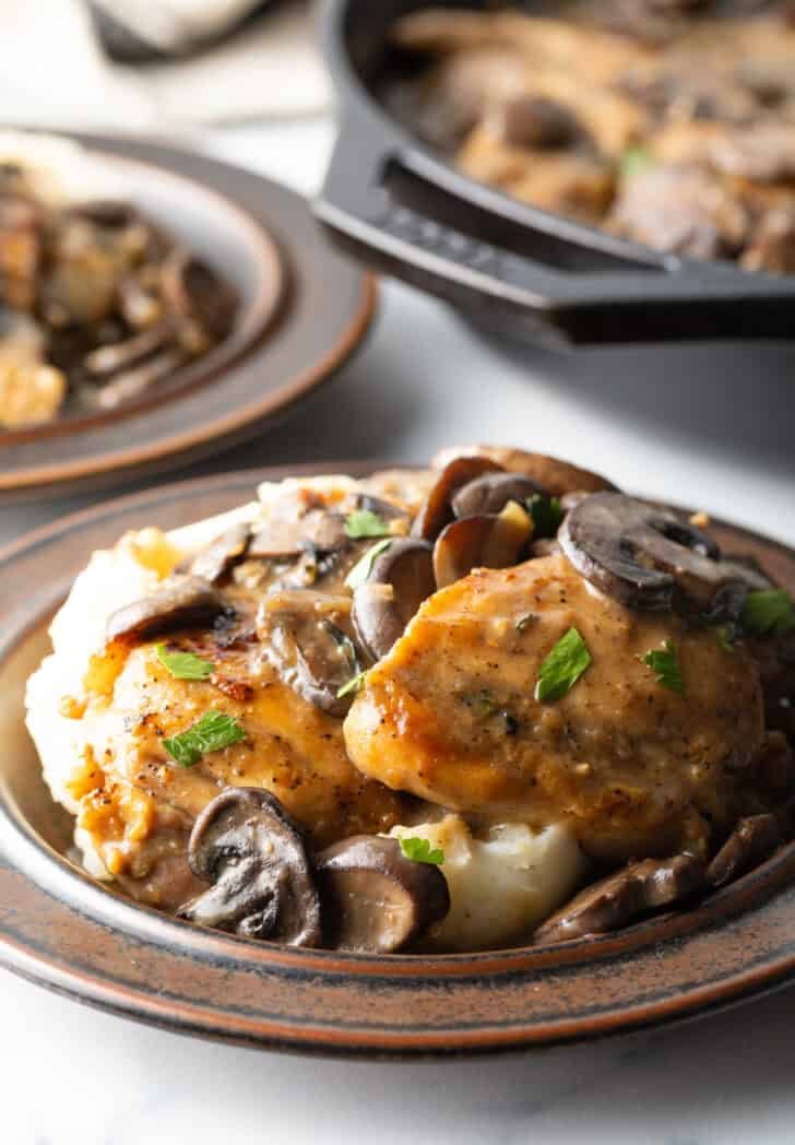 Brown plate with mashed potatoes, topped with two chicken breasts, sliced mushrooms, and gravy.