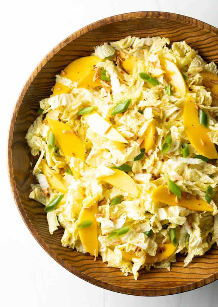 Top down view wood bowl with green napa cabbage, mango slices, and green onions.