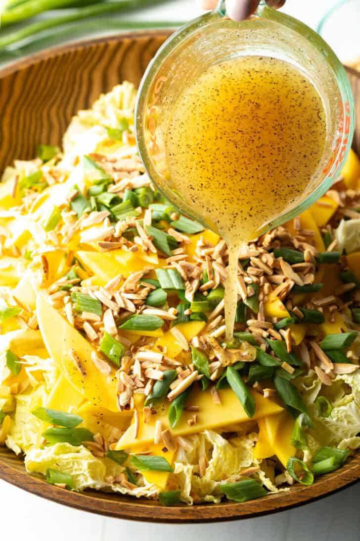Pouring dressing from a glass measuring cup into a bowl with shredded cabbage, toasted almonds, green onion slices, and slices of mango.