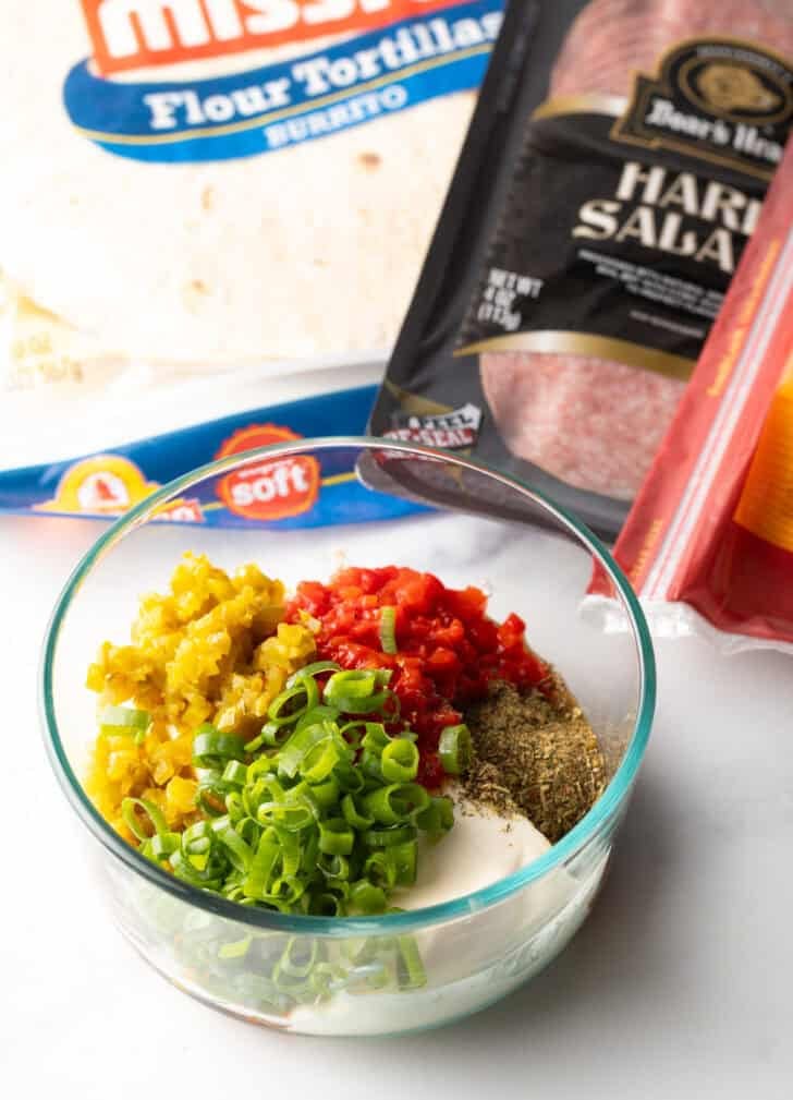 Glass bowl with pinwheel filling ingredients: chopped green onions, cream cheese, red peppers, and jalapenos.