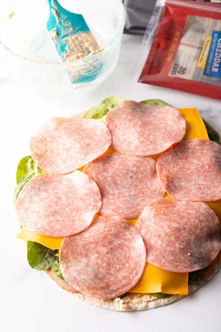 Slices of salami covering cheese and spinach on a tortilla.