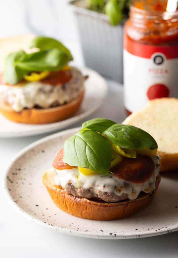 Open face burger on a white and black speckled plate. On the bottom bun is sauce, a stacked burger patty topped with melted mozzarella cheese, pepperoni slices, banana pepper rings, and fresh baby basil leaves.