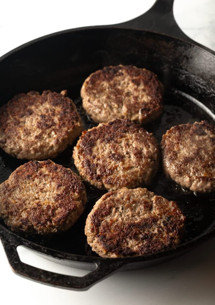 6 hamburgers cooking in a cast iron skillet.