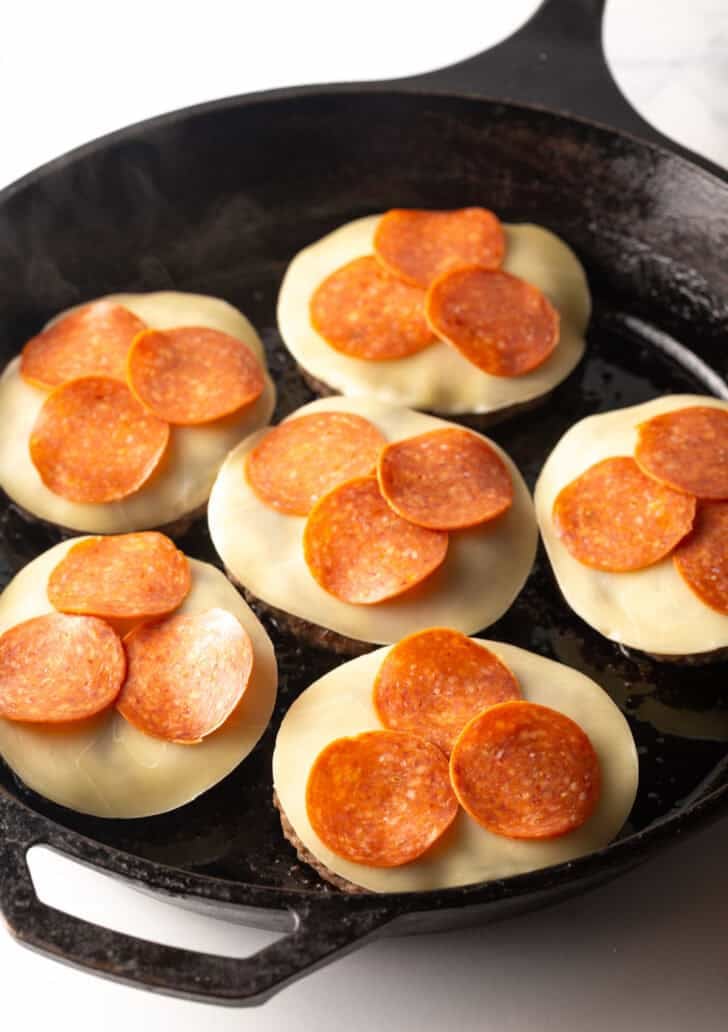 6 burger patties topped with mozzarella cheese slices and pepperoni slices in a cast iron skillet.