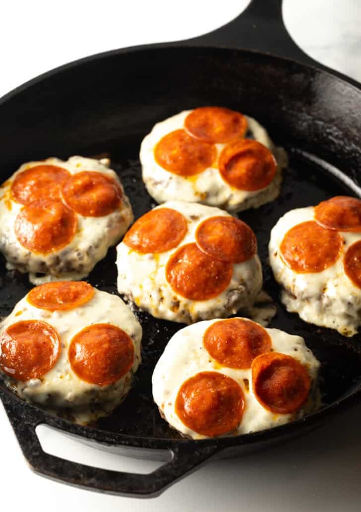 6 burger patties topped with melted mozzarella cheese and pepperoni slices in a cast iron skillet.