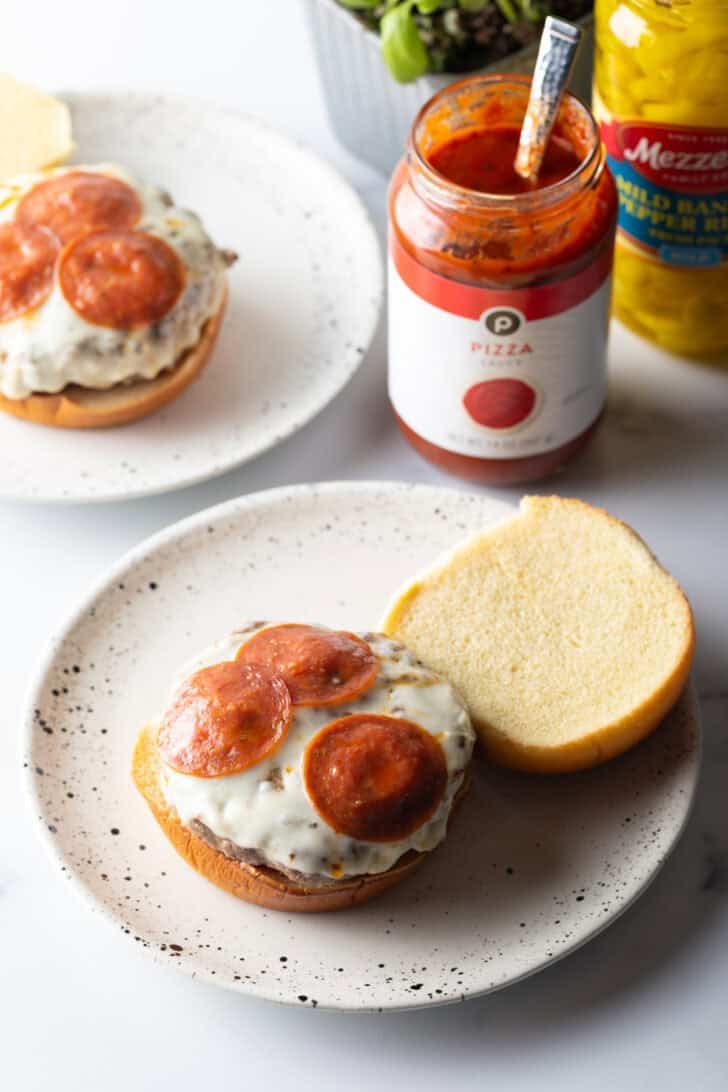Open face burger on a white and black speckled plate. On the bottom bun is sauce, a stacked burger patty topped with melted mozzarella cheese, and pepperoni slices.